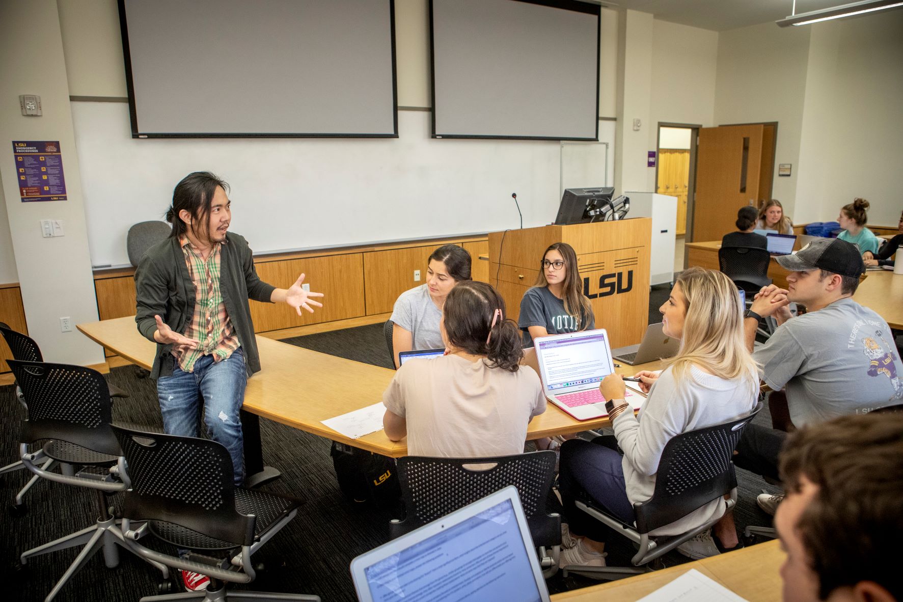 Man sits on desk and speaks while students look on. 